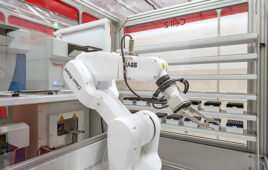 A white robotic arm with a two prong gripper at the end in a white and red lab.