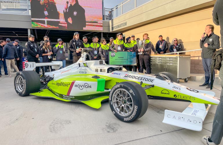 The Team Polimove team gathered around their vehicle in the Indy Autonomous Challenge winners circle.